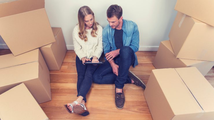 Young couple sitting in new house with packing boxes. They look like they have just moved in and they are very happy, smiling and talking. They are using a digital tablet, sitting on the floor in casual clothes. Copy space.